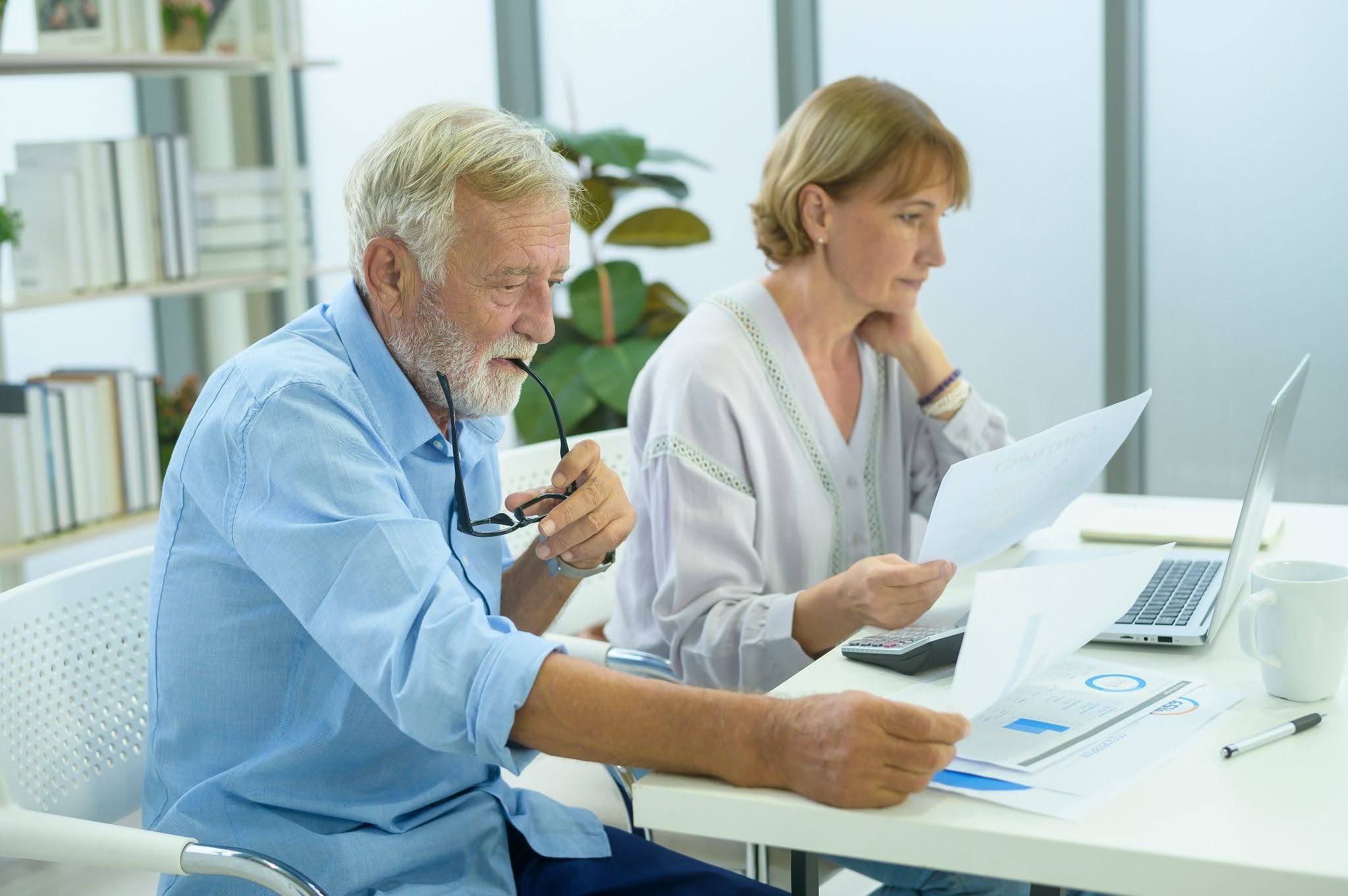 Senior couple reviewing retirement income documents at a desk with a laptop, calculator, and financial charts.