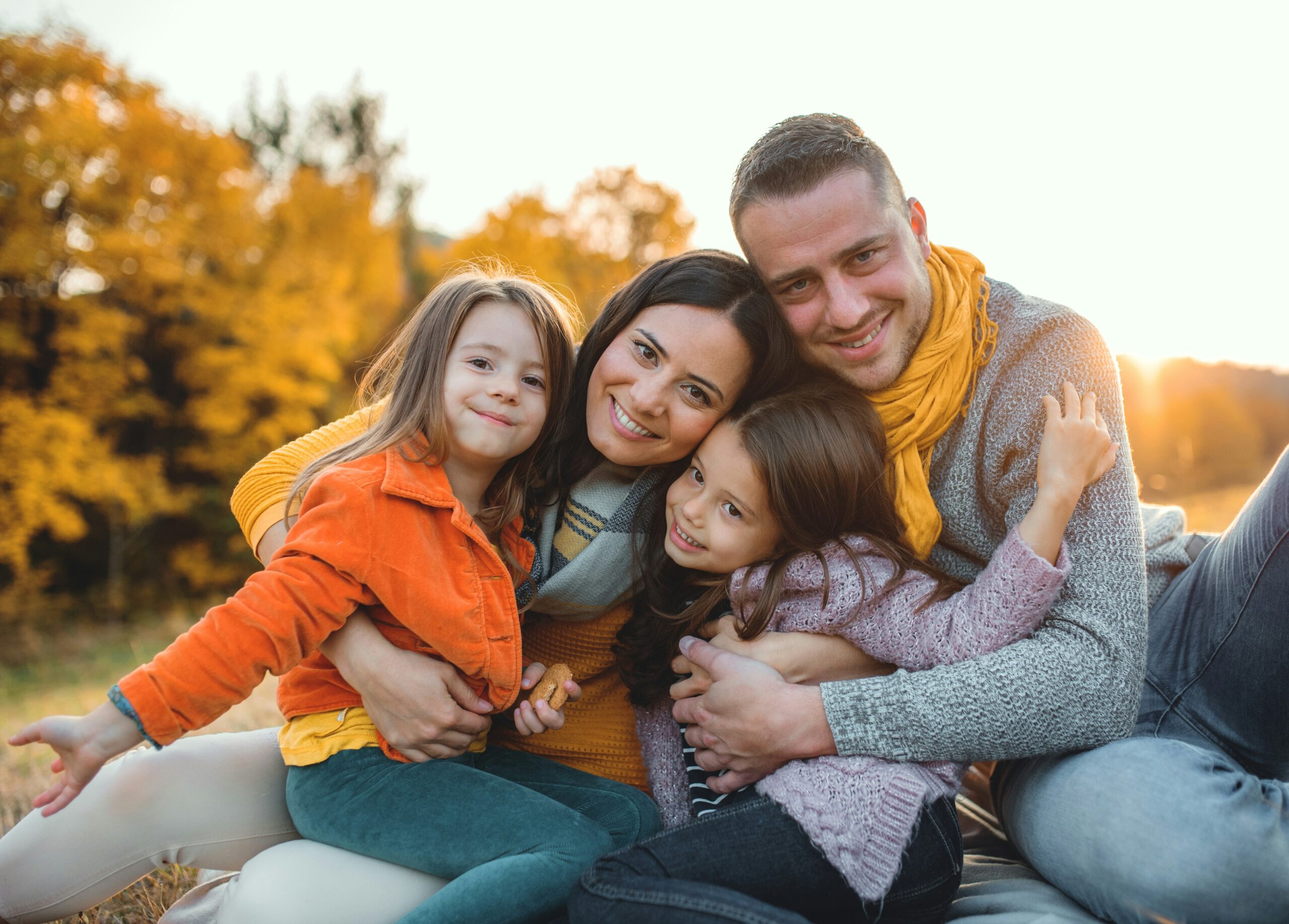 A smiling father, mother, and young daughters sitting close together outdoors in a sunny park.