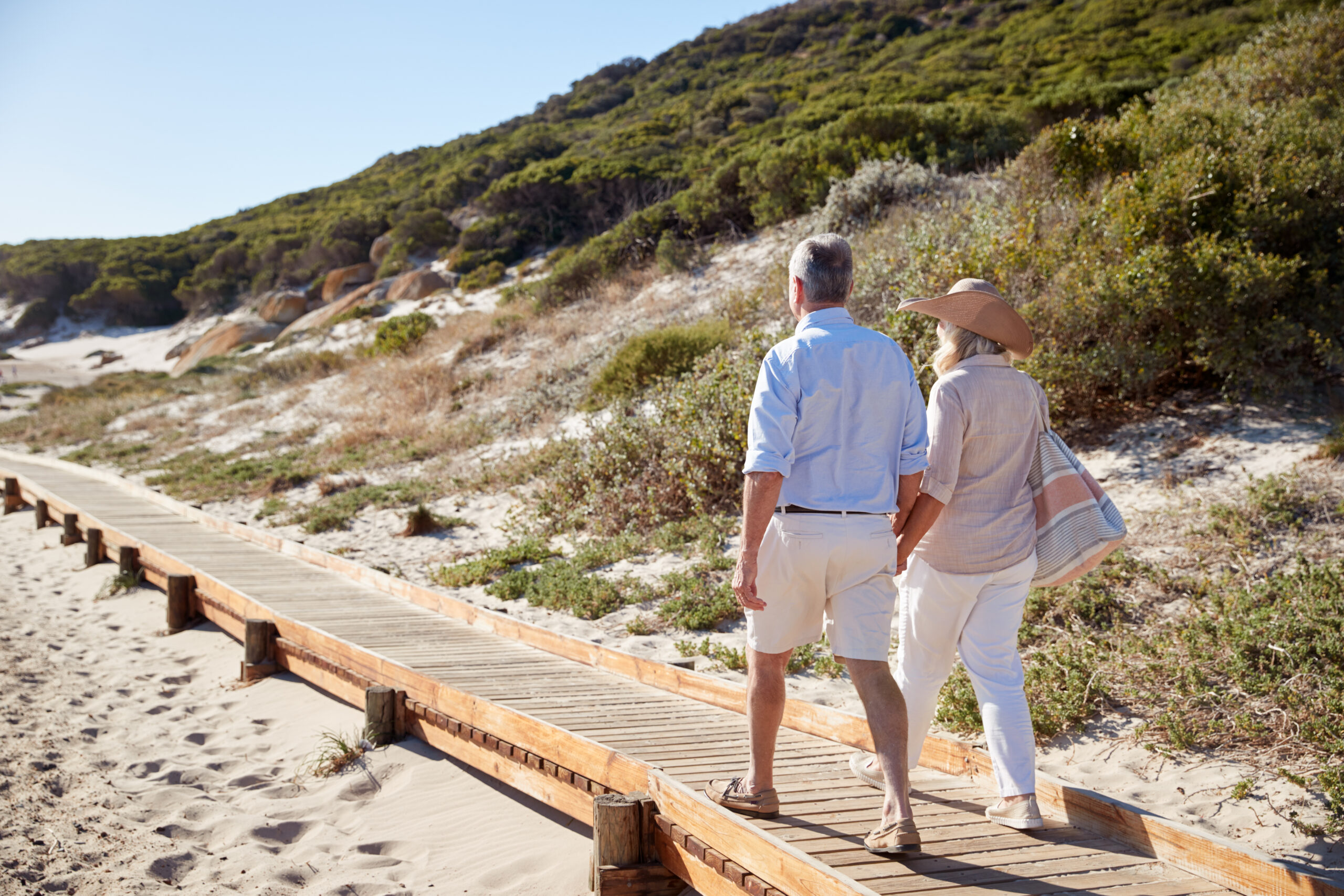 A man and woman walking down a boardwalk path framed by sand dunes and beach vegetation.